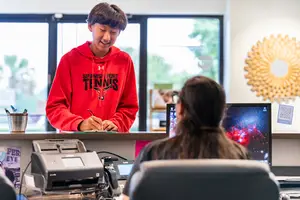 A boy in a red hoodie standing at a desk with a woman sitting in front of him looking at a computer screen
