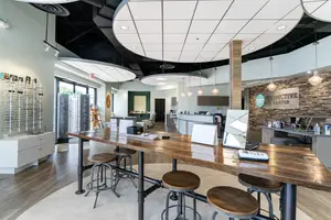 A wide shot of an office interior with a large table and stools, a wall with a stone design, and a glass window, all with a modern and clean look.