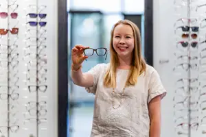 A smiling woman holds up a pair of glasses in a store with other glasses on display in the background