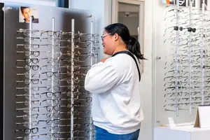 A woman with glasses stands in front of a display of eyeglasses in a store.