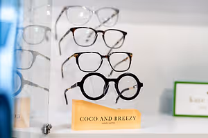 An array of eyeglasses displayed on a white shelf with a glass cover, including a pair with the label Coco and Breezy Handcrafted