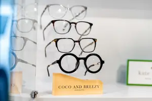An array of eyeglasses displayed on a white shelf with a glass cover, including a pair with the label Coco and Breezy Handcrafted