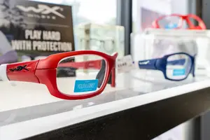 Several pairs of sport glasses are displayed on a white shelf, including a red pair with a blue label that says Youth Force and a blue pair in a transparent container.
