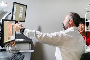 A man in a lab coat points at an image on a computer screen while another man watches behind him.