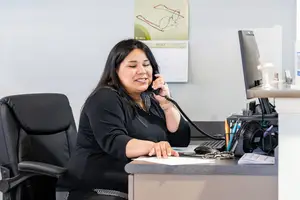 A smiling woman wearing a black top is sitting at a desk, holding a phone to her ear and seems to be talking to someone. There is a monitor, a keyboard, a mouse, and a pen holder with pens on the desk. Behind her is a wall with a calendar and a drawing of eyeglasses.