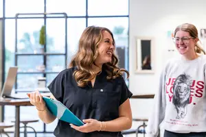 Two women standing and smiling in a room with a glass window in the background