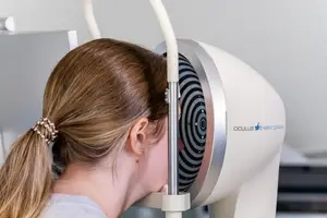 A woman with blonde hair wearing a gray shirt is using an eye examination machine in a white room.