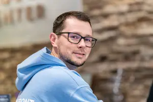 Closeup of a man wearing a light blue hoodie and black glasses standing in front of a wall