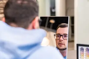 A man in a blue hoodie is looking at himself in a mirror while wearing glasses.