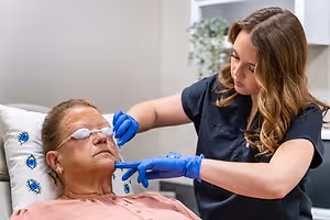 An older woman is receiving eye treatment from a woman in a blue shirt in a medical setting.
