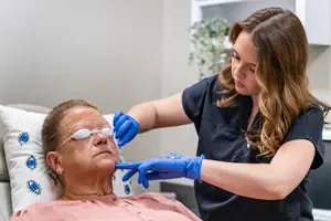 An older woman is receiving eye treatment from a woman in a blue shirt in a medical setting.