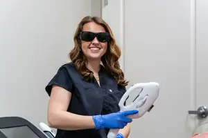 A woman wearing sunglasses and blue gloves holds a white medical laser device in a room.