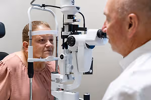 A woman is undergoing an eye examination with a man standing next to her using an ophthalmoscope