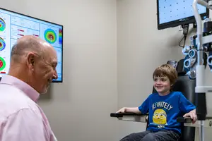 A young boy is sitting in a chair in front of a doctor who is looking at a monitor with eye charts and diagrams, while the boy is smiling and looking at the camera.