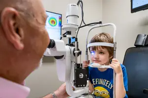 A young boy with glasses is having an eye exam by an older man in a medical setting.