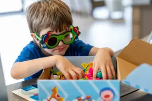 A young boy wearing parrot-themed sunglasses is sorting colorful toys into a box.