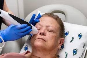 A woman lying on a bed is having her eyebrows groomed by a beautician wearing gloves.
