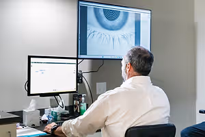 A man in a white shirt is sitting at a desk and looking at a monitor with an eye image on it.