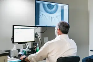 A man in a white shirt is sitting at a desk and looking at a monitor with an eye image on it.