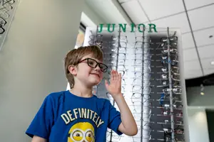 A smiling boy in eyeglasses waves in front of a display of eyeglasses at a Junior Eyewear store.