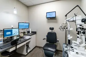A small room with a desk, monitor, and a black chair next to an eye examination machine, mounted monitor, and sink.