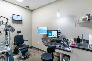 A medical room with a chair, desk, computer monitor, sink, and other equipment