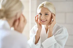 A woman in a white robe is looking at her reflection in a mirror while smiling