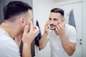 A man in a white t-shirt is looking at himself in the mirror, possibly checking his appearance.