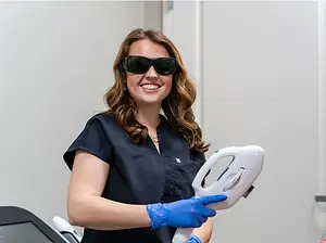 A woman wearing a black shirt and blue gloves holds a white medical laser device, likely for a skincare treatment