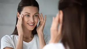 A woman is smiling and looking at her reflection in a mirror, with her hands on her face.