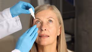 A doctor is checking the eyes of a woman who is sitting inside a room. She has blue eyes, a nose, and a mouth. The doctor is wearing a white coat and blue gloves. The woman is looking at the doctor.
