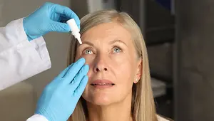 A doctor is checking the eyes of a woman who is sitting inside a room. She has blue eyes, a nose, and a mouth. The doctor is wearing a white coat and blue gloves. The woman is looking at the doctor.