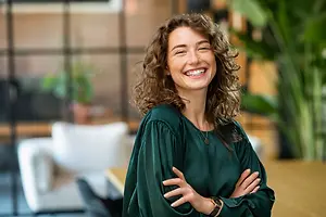A smiling woman with curly hair stands in an office with a desk and a white couch in the background.