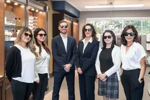 A group of five people, including three women and two men, are posing for a photo while wearing sunglasses in an indoor area, possibly a store, with wooden shelves and a glass window behind them.