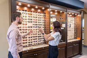 A man and a woman standing in front of a cabinet filled with sunglasses in a shop