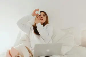 A woman sitting on a bed in her bedroom applying eye drops.