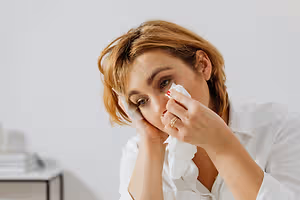 A woman with short hair is blowing her nose with a tissue while looking down.