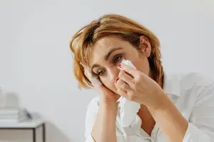 A woman with short hair is blowing her nose with a tissue while looking down.