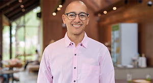 A man is smiling in a restaurant with a refrigerator and a table in the background.