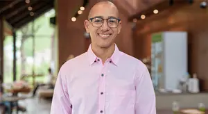 A man is smiling in a restaurant with a refrigerator and a table in the background.
