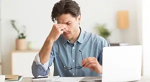 A man sitting at a desk rubbing his eyes and looking stressed