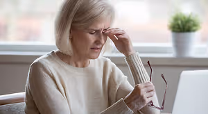 An elderly woman with short blonde hair is sitting on a couch, looking down with a worried expression on her face. She is holding a pair of glasses and her left hand is touching her forehead. There is a laptop on the table in front of her, and a potted plant is on the window sill behind her.