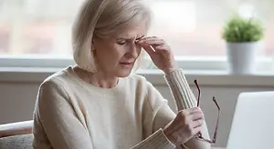 An elderly woman with short blonde hair is sitting on a couch, looking down with a worried expression on her face. She is holding a pair of glasses and her left hand is touching her forehead. There is a laptop on the table in front of her, and a potted plant is on the window sill behind her.