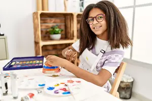 A young girl wearing glasses and an apron is painting with a paint palette on a table.