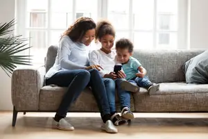 A family of three, a woman and two children, are sitting on a couch and looking at a cellphone.