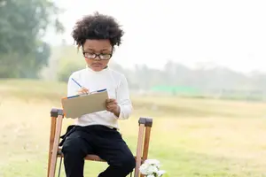 A young boy is sitting in a chair in a park, holding a book and a pen, and is wearing glasses.