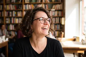 A smiling woman in glasses sits in a library surrounded by bookshelves, with another person sitting at a table in the background.