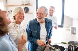 A group of older adults are smiling and laughing in the kitchen, with one man holding a wooden spoon and the other holding a glass of wine.