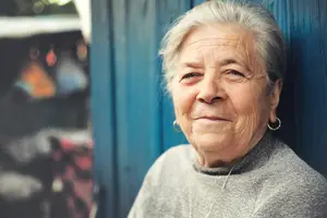An elderly woman with white hair and a gray sweater leaning against a blue wall, with a blurry background of a motorcycle and other objects.