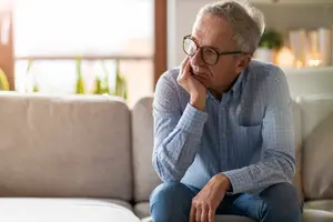 An elderly man is sitting on a couch with his hand on his face, looking sad.